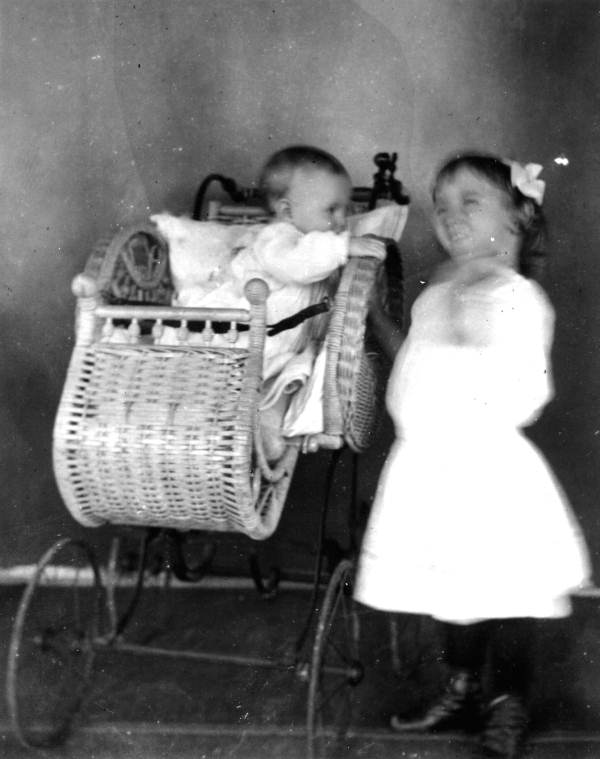 Little girl standing by infant in perambulator.