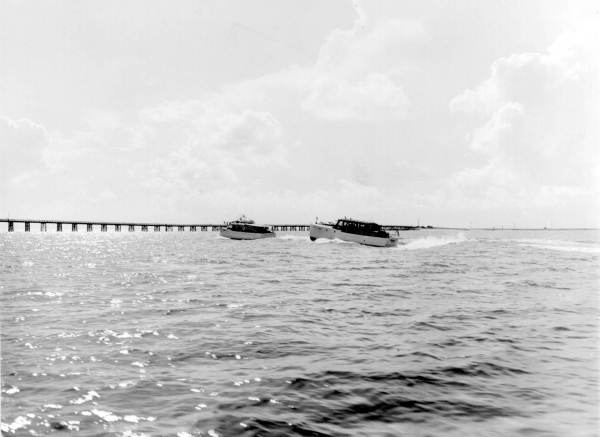Two small yachts glide through the water with a pier in the background
