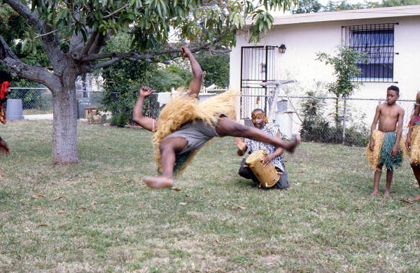 Nigerian acrobatic dancer performs as apprentice Moses Campbell drums - North Miami, Florida.
