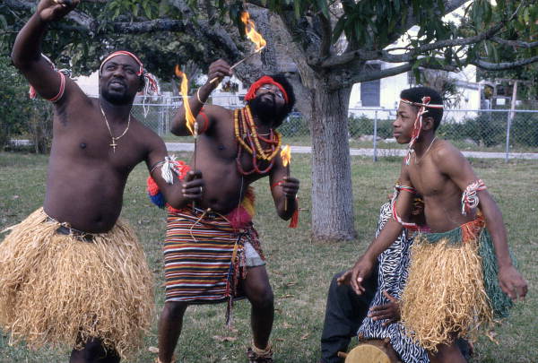 Nigerian acrobatic dancers Augustine and Felix Omeben preparing to eat fire while apprentice Akila Baki observes - North Miami, Florida.