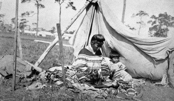 Seminole women sewing under canvas shelter with child beside her - Florida