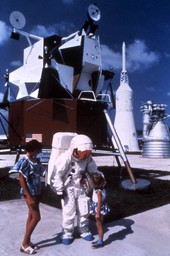 Mother and daughter being greeted by person in space suit next to lunar module on display in the Kennedy Space Center rocket garden.